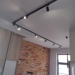 Czarny system szynowy z reflektorami punktowymi zamontowany na suficie w pomieszczeniu z ceglaną ścianą i częściowo zamontowaną zabudową meblową; w tle drabina i osoba w niebieskiej koszulce.