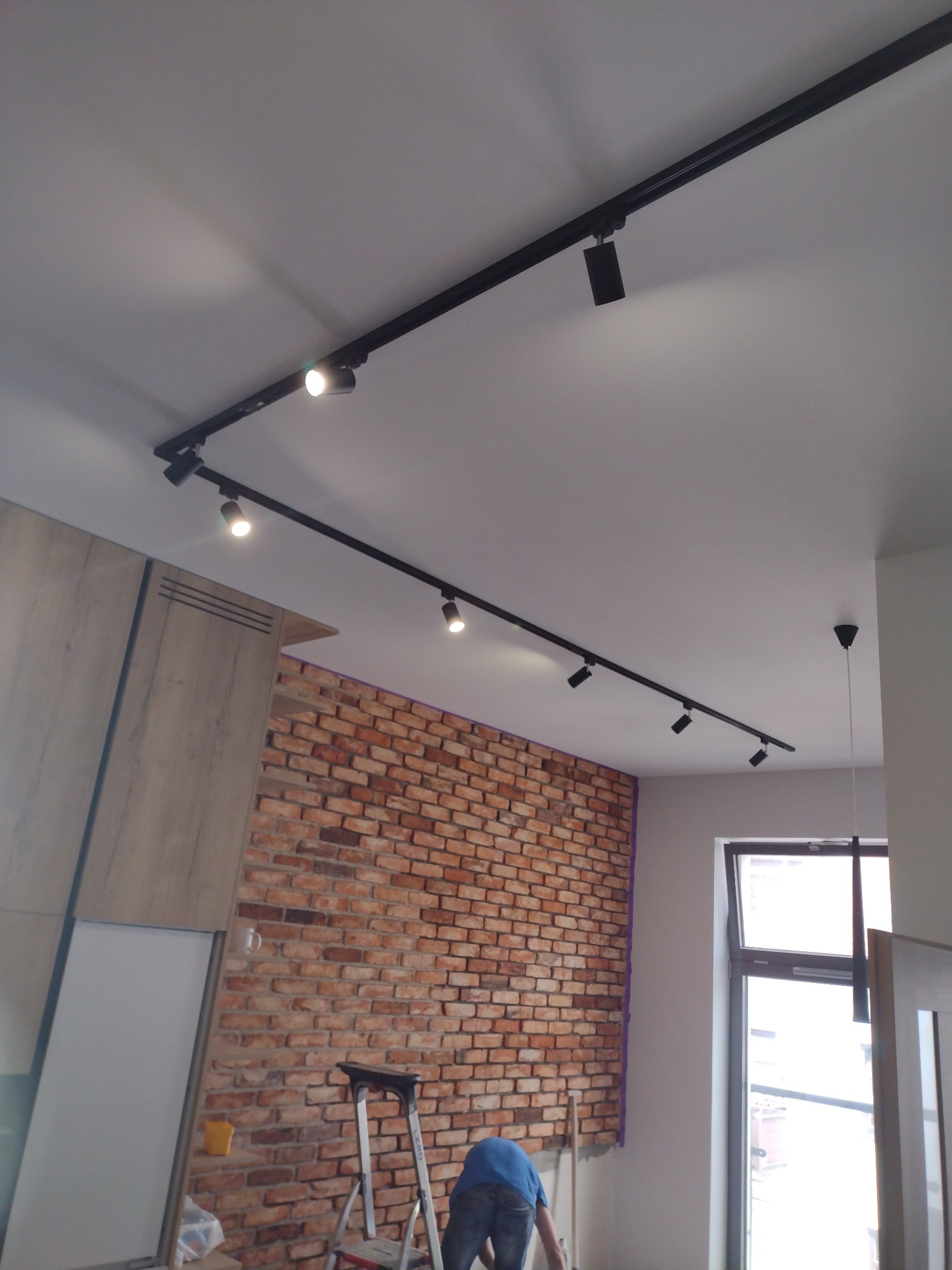 Czarny system szynowy z reflektorami punktowymi zamontowany na suficie w pomieszczeniu z ceglaną ścianą i częściowo zamontowaną zabudową meblową; w tle drabina i osoba w niebieskiej koszulce.