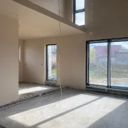 Wnętrze domu w trakcie prac wykończeniowych, ściany pokryte świeżym, jasnym tynkiem, widoczne kable elektryczne wystające ze ścian i sufitu, duże okna z widokiem na zewnątrz, podłoga w stanie...