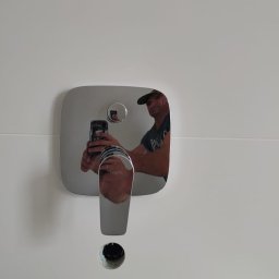 Cover AT bud Andrii Danyliuk - Bateria umywalkowa wmontowana w białą ścianę z widocznym otworem na odpływ i odbiciem instalatora robiącego zdjęcie smartfonem.