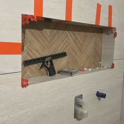 Cover AT bud Andrii Danyliuk - Narożnik wnęki wyłożony płytkami w jodełkę, zabezpieczony pomarańczową taśmą i poziomowany. Widoczne narzędzia glazurnicze i fragment ściany z instalacją.