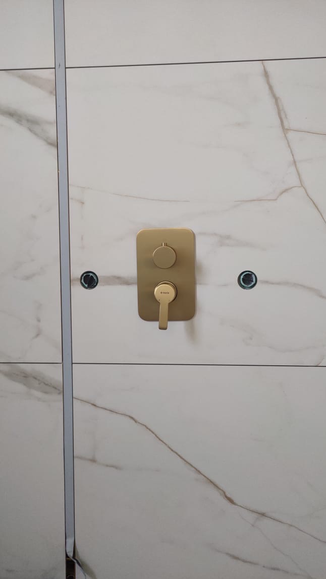 Ściana łazienki wyłożona płytkami imitującymi marmur z zamontowaną złotą baterią podtynkową i widocznymi otworami na inne elementy armatury.