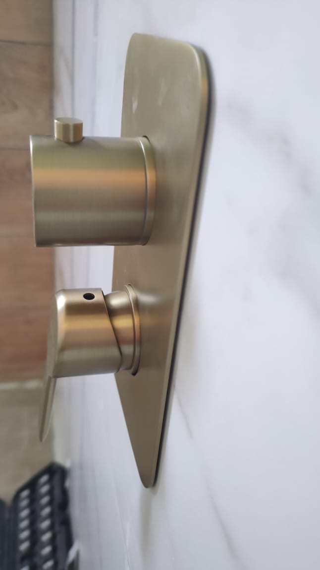 Bateria prysznicowa w odcieniach złota, zamontowana na białej, marmurowej ścianie w łazience. Widoczny fragment drewnopodobnej okładziny ściany oraz czarna skrzynka narzędziowa.