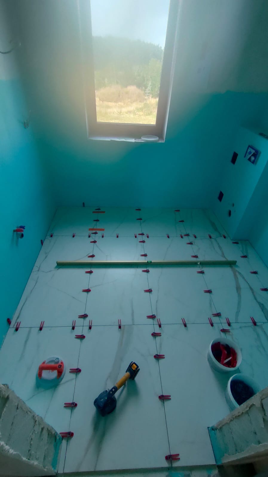 Ściana w łazience na poddaszu z oknem dachowym, w trakcie układania białych płytek z marmurkowym wzorem, używane czerwone klipsy poziomujące i przyssawka do płytek.