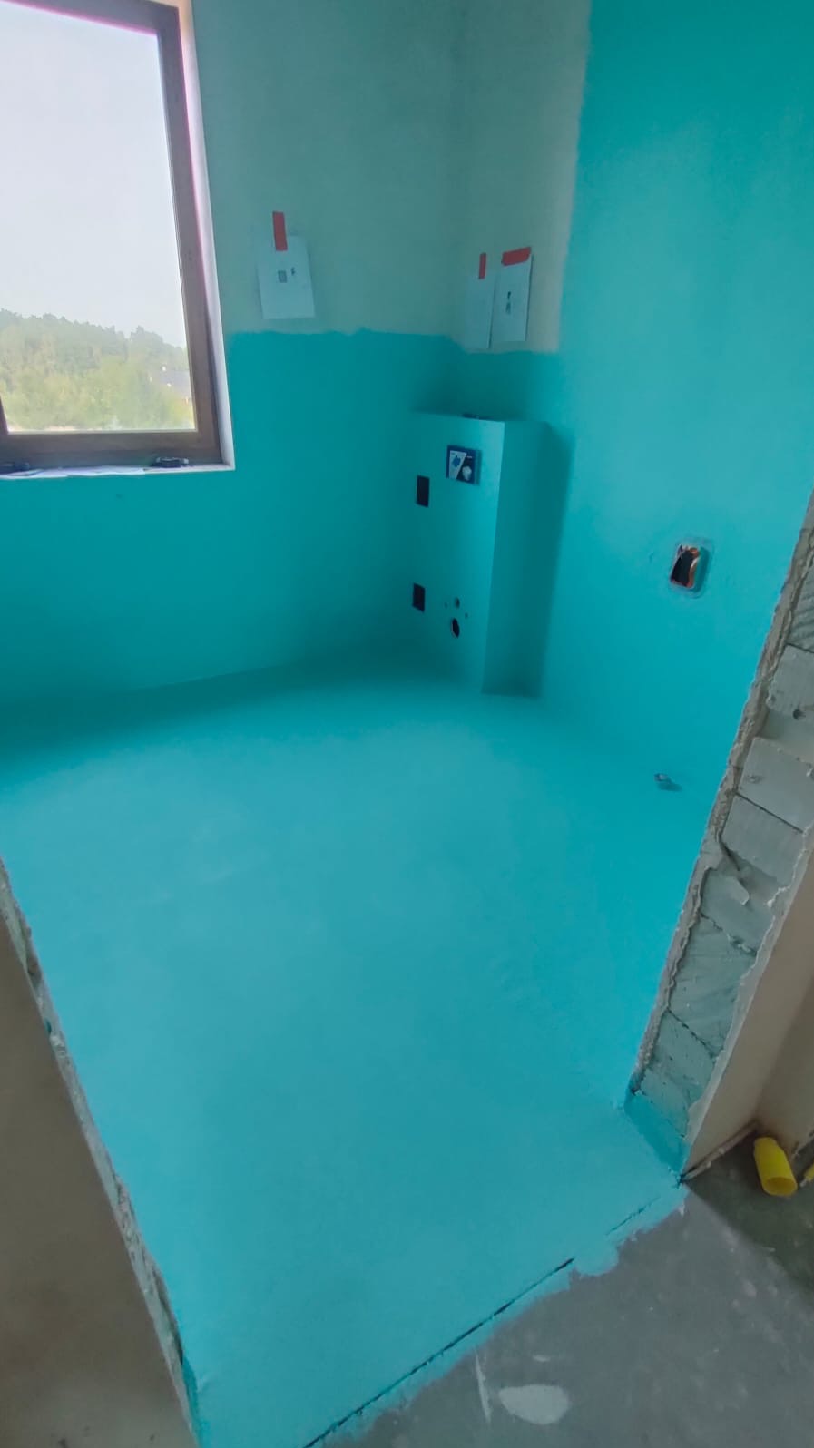 Niebieska hydroizolacja w łazience w trakcie remontu, widoczne przygotowane podejścia pod instalacje elektryczne i wodne, fragment okna i surowy fragment ściany z widocznymi cegłami.