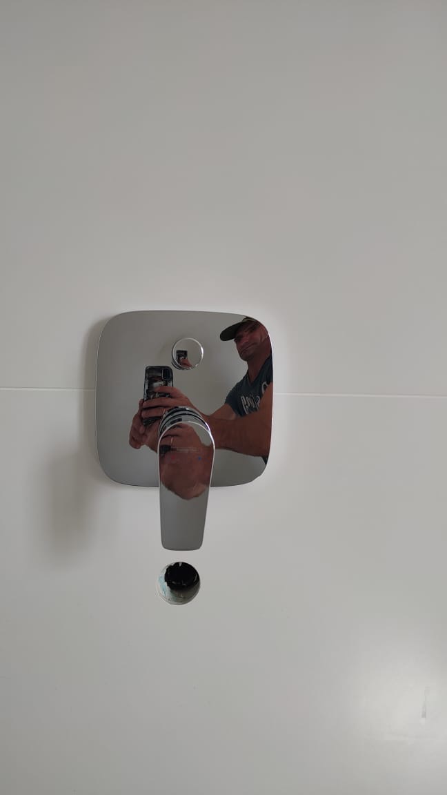 Bateria umywalkowa wmontowana w białą ścianę z widocznym otworem na odpływ i odbiciem instalatora robiącego zdjęcie smartfonem.