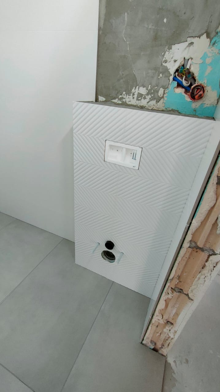 Stelaż podtynkowy WC zabudowany płytkami z delikatnym wzorem chevron, widoczne przyłącza wody i odpływu, fragment surowej ściany z instalacją elektryczną w tle.