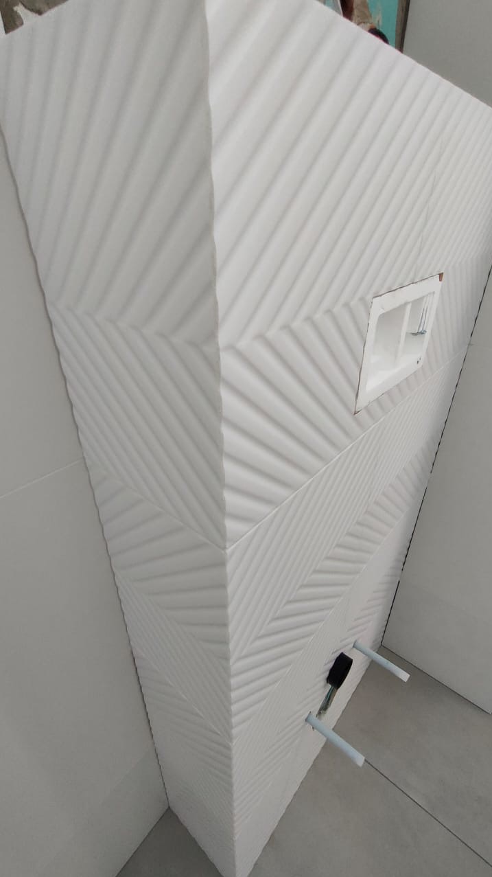 Nowoczesna, biała ściana w łazience wyłożona trójwymiarowymi płytkami ceramicznymi z wzorem chevron, widoczne elementy instalacji sanitarnej i podtynkowy stelaż WC.