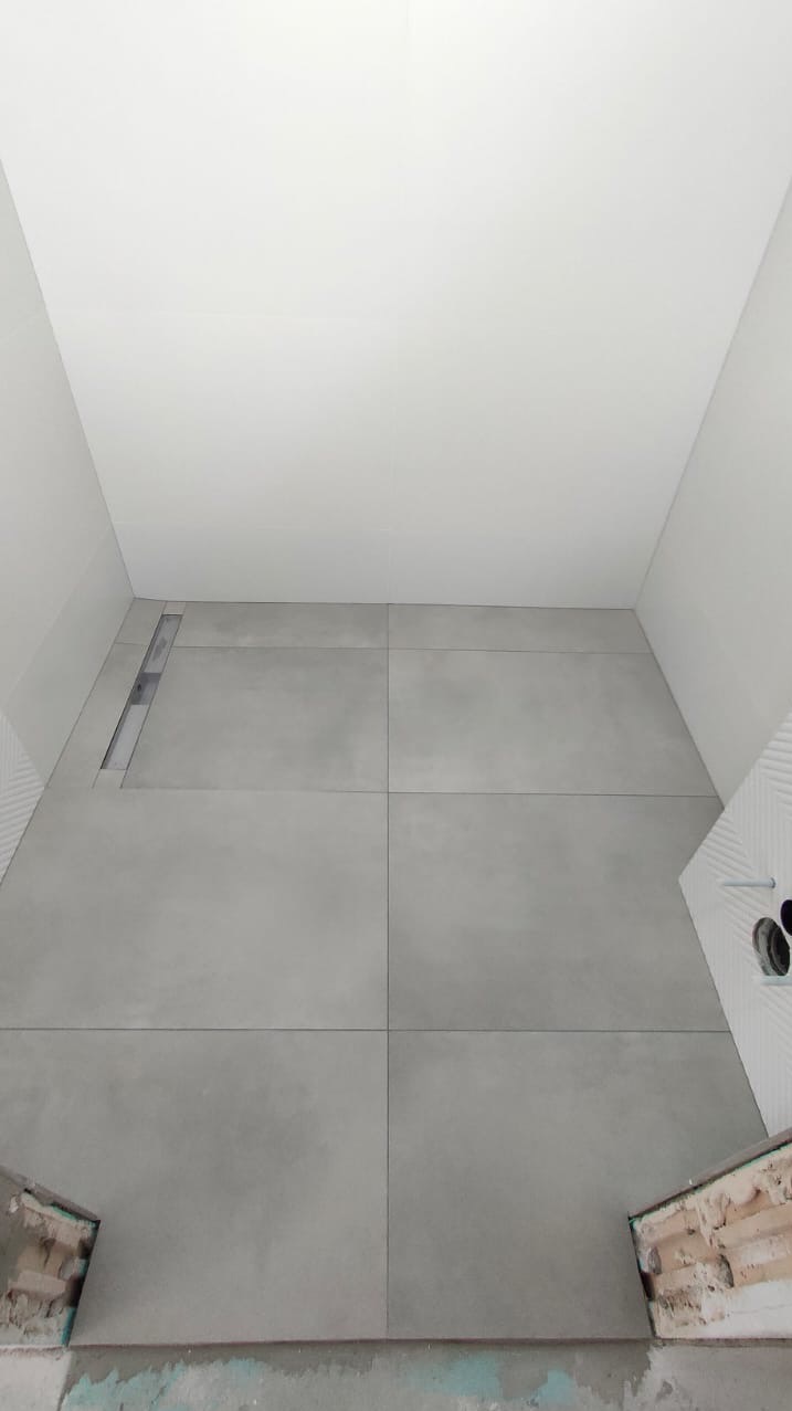 Szara, minimalistyczna podłoga z dużych płytek w łazience w Szczecinie, z widocznym odpływem liniowym i fragmentem ściany z dekoracyjnymi panelami.