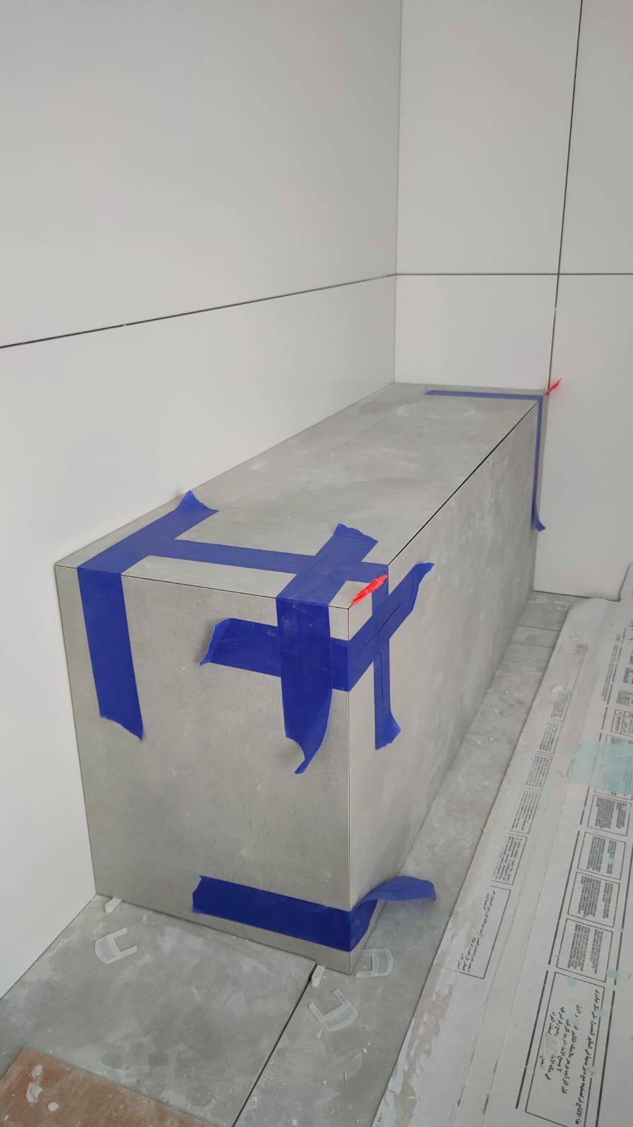 Wnętrze łazienki w trakcie remontu: obłożona płytkami ściana i obudowa wanny lub brodzika, zabezpieczona niebieską taśmą malarską, z widocznymi krzyżykami dystansowymi do płytek na podłodze.