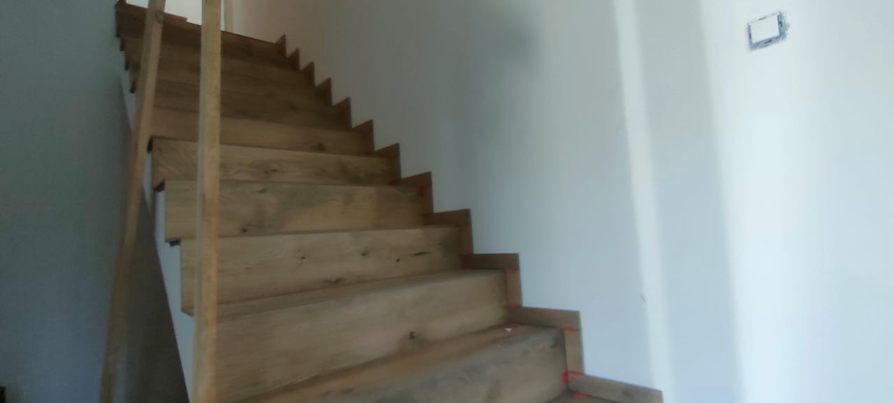 Drewniane schody w jasnym odcieniu, widoczne z dołu, z surowymi ścianami i wystającym gniazdkiem elektrycznym w tle. Realizacja w trakcie wykończenia.