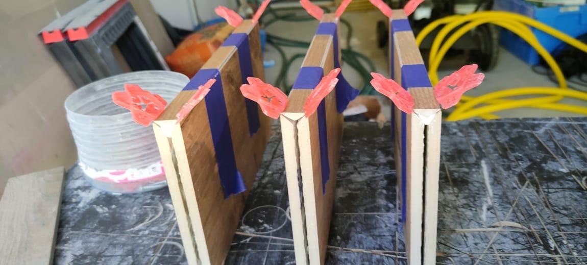 Trzy drewniane panele z niebieską taśmą i czerwonymi klamrami, ustawione pionowo na blacie, ukazujące proces budowy lub naprawy.