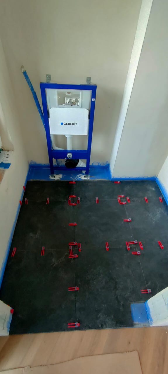 Montaż stelaża podtynkowego Geberit i układanie ciemnych płytek podłogowych z czerwonymi krzyżykami dystansowymi w łazience, zabezpieczone niebieską folią.