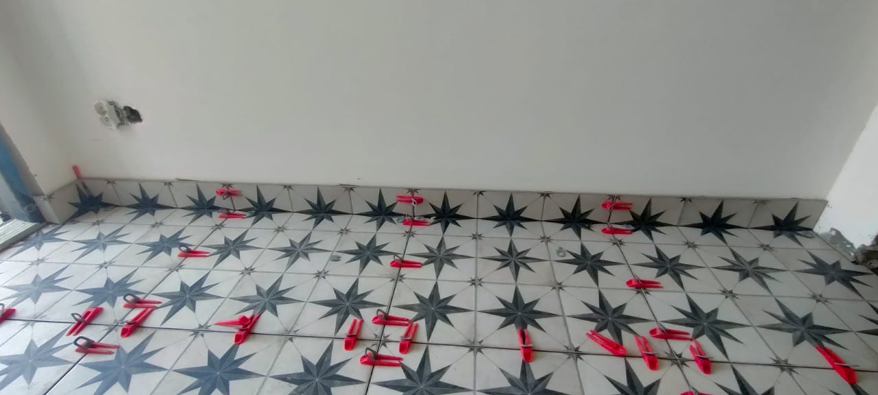 Układanie płytek podłogowych w gwiazdy z użyciem czerwonych klipsów poziomujących. Widoczny fragment ściany i niedokończone wykończenie.