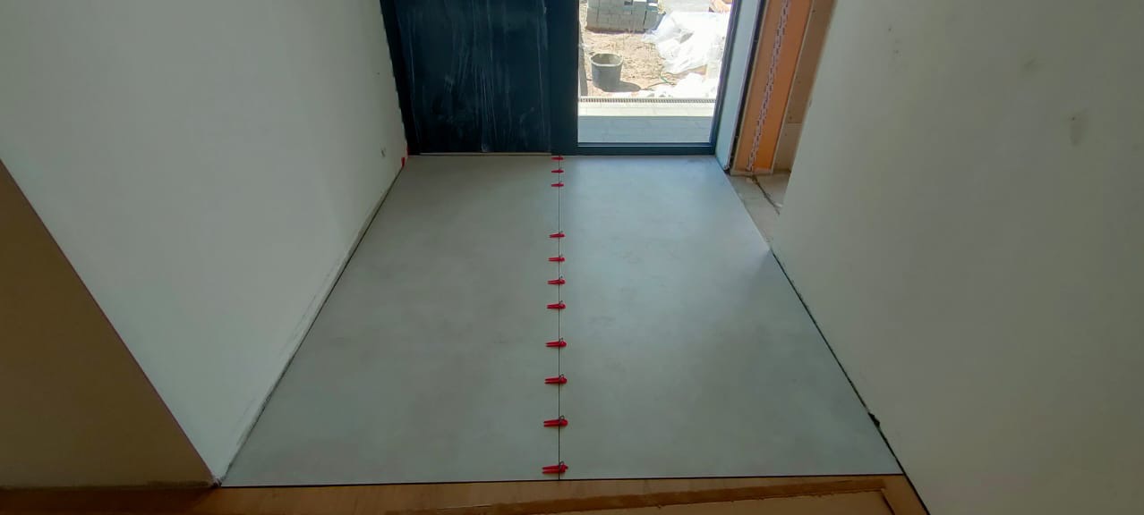 Ułożone płytki podłogowe w korytarzu, z użyciem czerwonych klipsów poziomujących. Widoczne gładkie, szare płytki i fragment ramy okiennej.