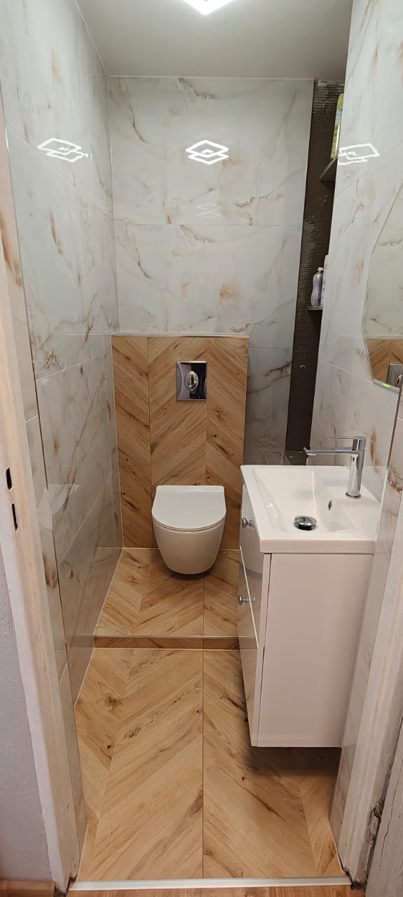 Nowoczesna łazienka z beżowymi płytkami imitującymi marmur i drewnopodobną podłogą ułożoną w jodełkę. Umywalka z szafką i podwieszana toaleta.