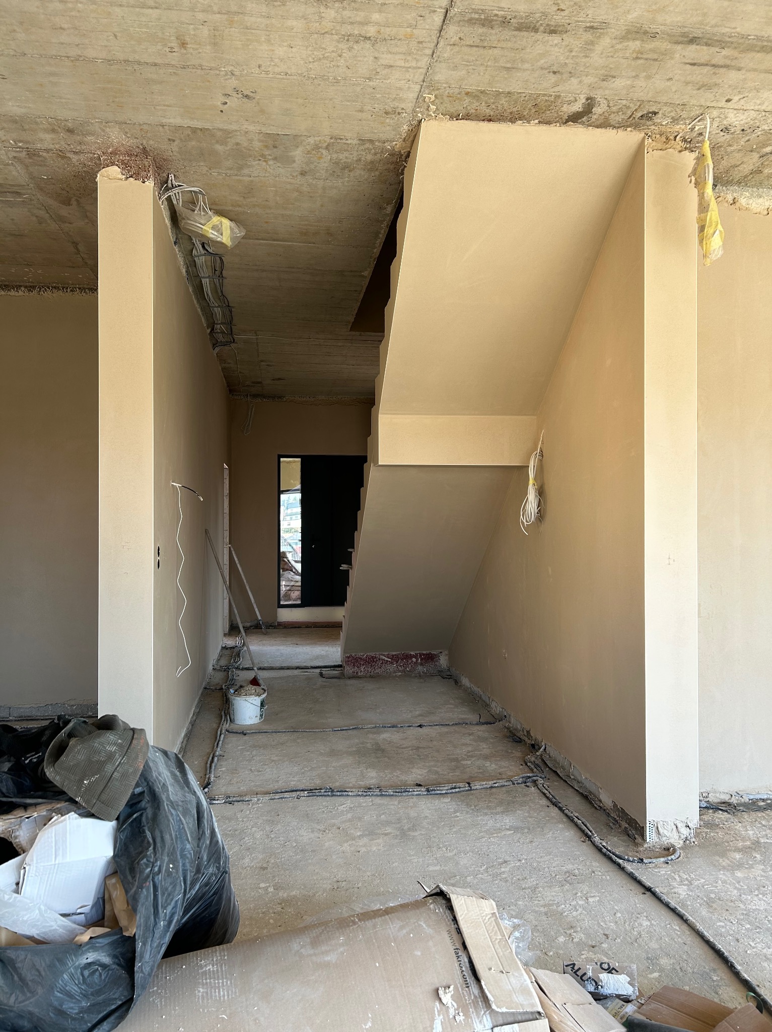 Wnętrze w trakcie prac tynkarskich: świeżo otynkowane ściany i schody w beżowym odcieniu, widoczne przewody elektryczne, surowy betonowy sufit i elementy wyposażenia budowlanego.