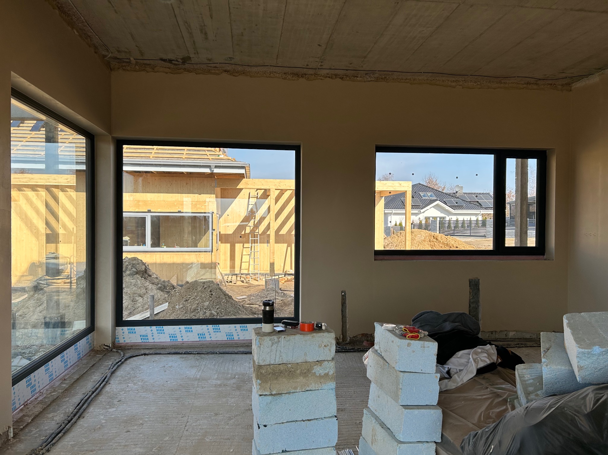Surowy stan wnętrza domu w budowie: widoczne okna z czarnymi ramami, ściany w kolorze beżowym, betonowy sufit, podłoga z wylewką i materiały budowlane.