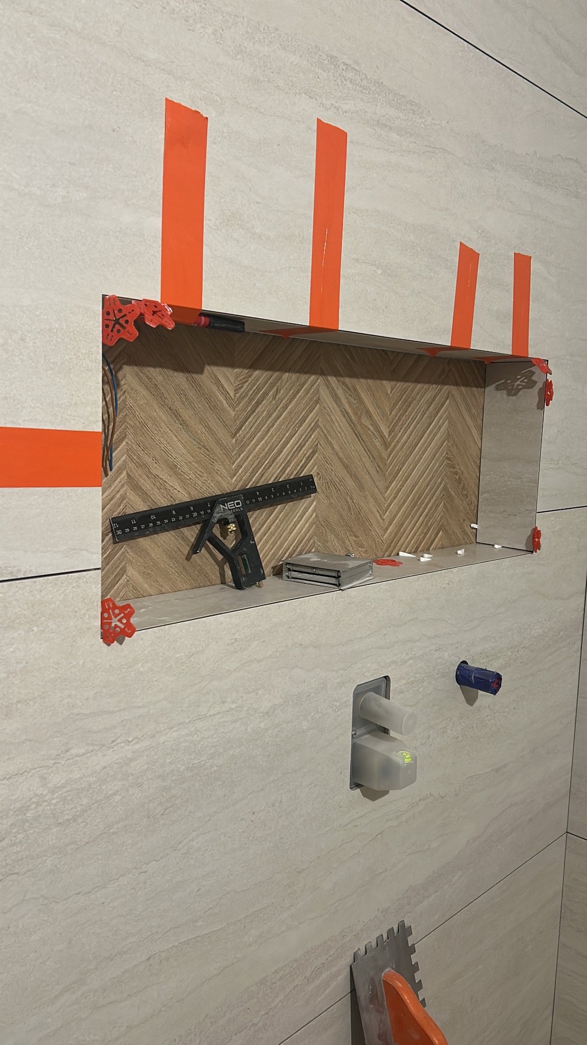 Narożnik wnęki wyłożony płytkami w jodełkę, zabezpieczony pomarańczową taśmą i poziomowany. Widoczne narzędzia glazurnicze i fragment ściany z instalacją.