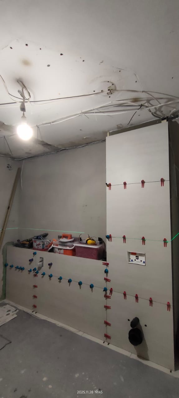 Ściana z płytkami w trakcie układania, widoczne krzyżyki dystansowe i poziomica laserowa. Prace wykończeniowe w łazience, surowy stan, widoczne przewody na suficie.