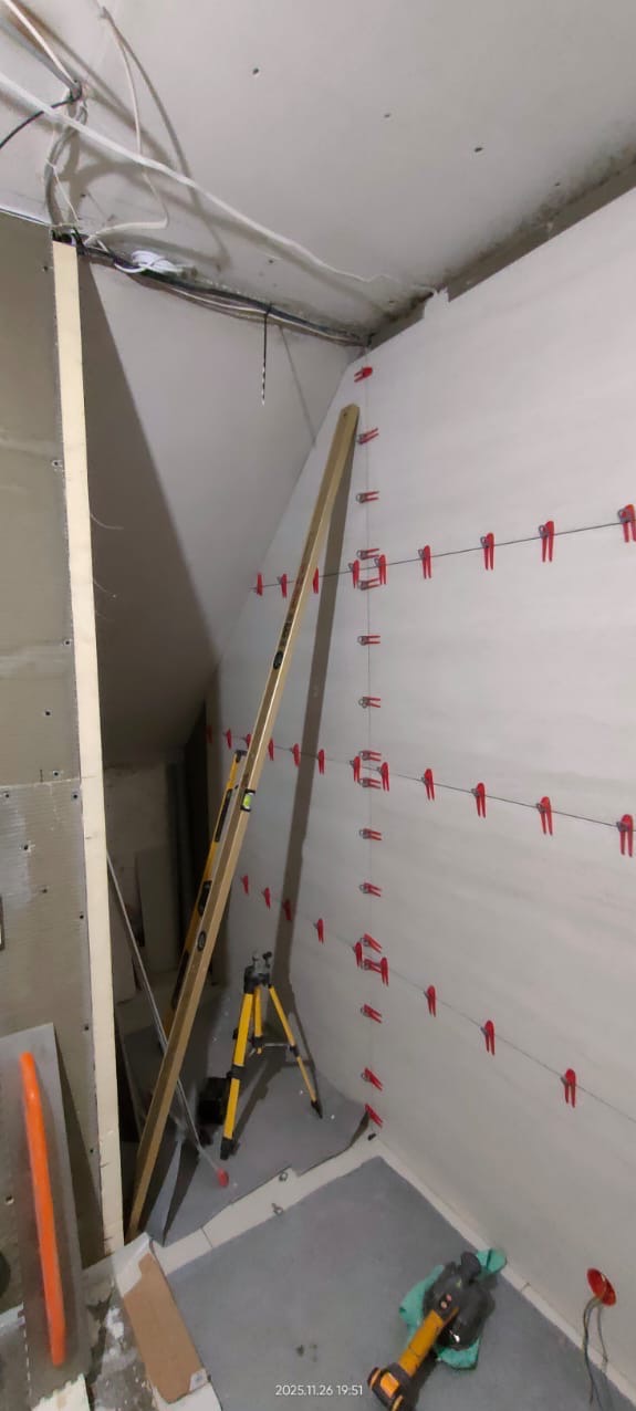 Ściana z poziomowaniem laserowym i klipsami do płytek w trakcie remontu. Widoczny poziomica, statyw i narzędzia. Prace glazurnicze w łazience ze skosem.