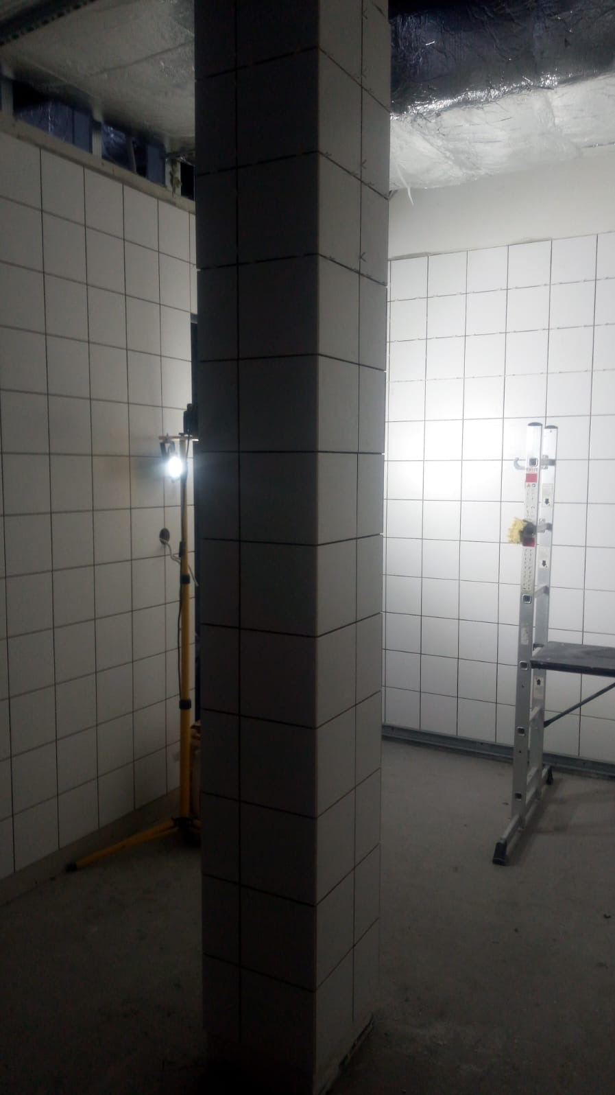 Wykończona kolumna i ściany w płytkach w surowym wnętrzu. Widoczna drabina i lampa budowlana. Minimalistyczny design z kwadratowymi płytkami w jasnym odcieniu szarości.