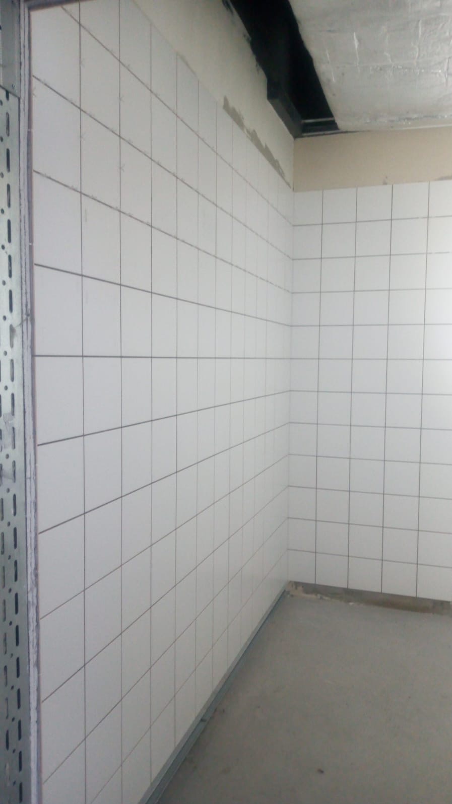 Wykończona ściana z białych, kwadratowych płytek w pomieszczeniu z szarą podłogą. Widoczna metalowa listwa przypodłogowa i fragment instalacji pod sufitem.