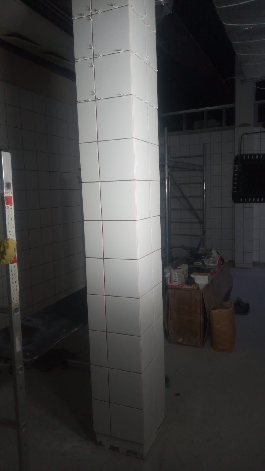 Biała kolumna w trakcie wykańczania, obłożona kwadratowymi płytkami z krzyżykami dystansowymi. W tle widoczne rusztowanie i inne elementy wyposażenia.