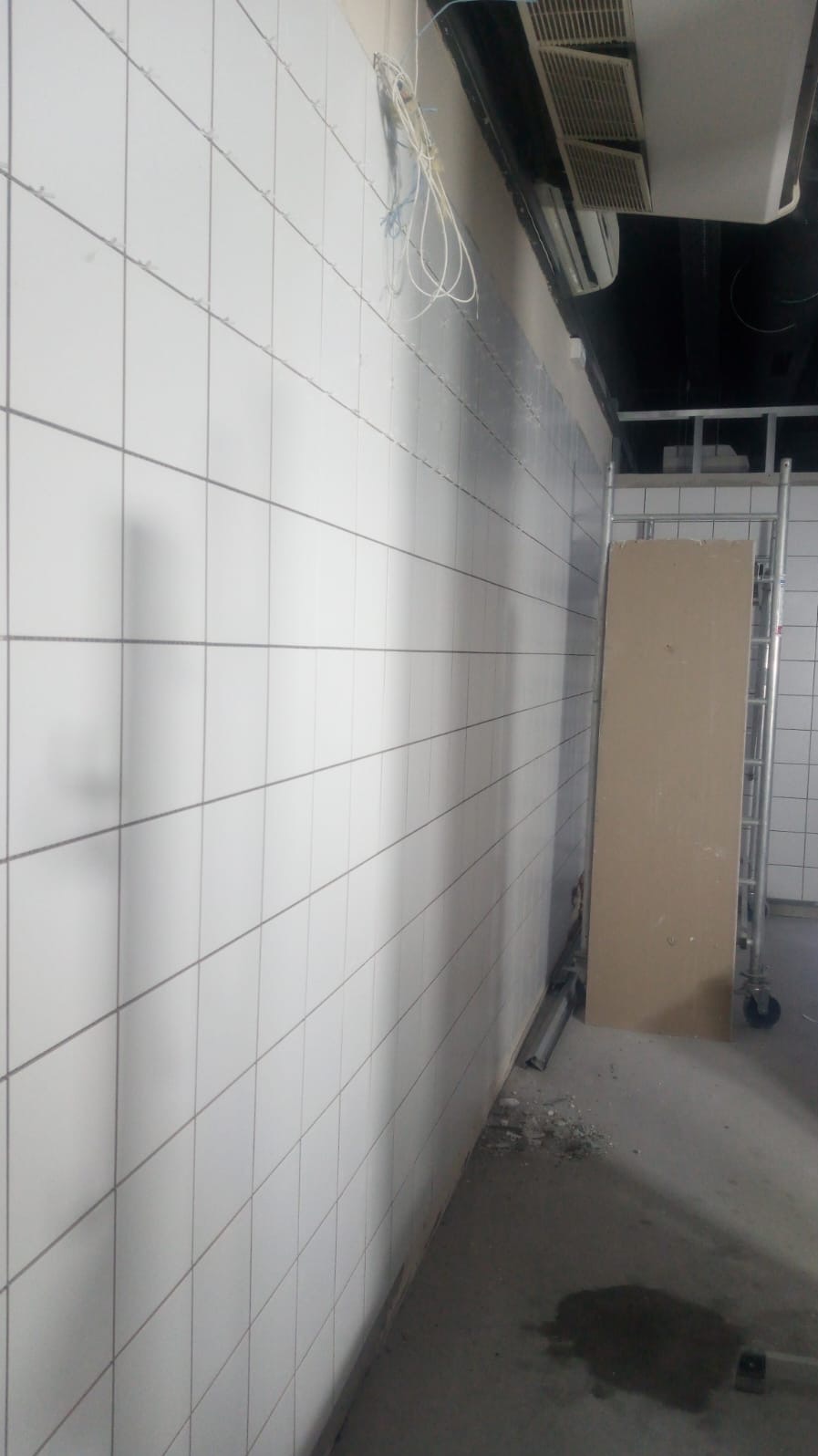 Białe, kwadratowe płytki na ścianie, widoczne przewody i fragment sufitu z klimatyzacją. Na podłodze resztki gruzu i plama wody. W tle metalowy regał na kółkach.