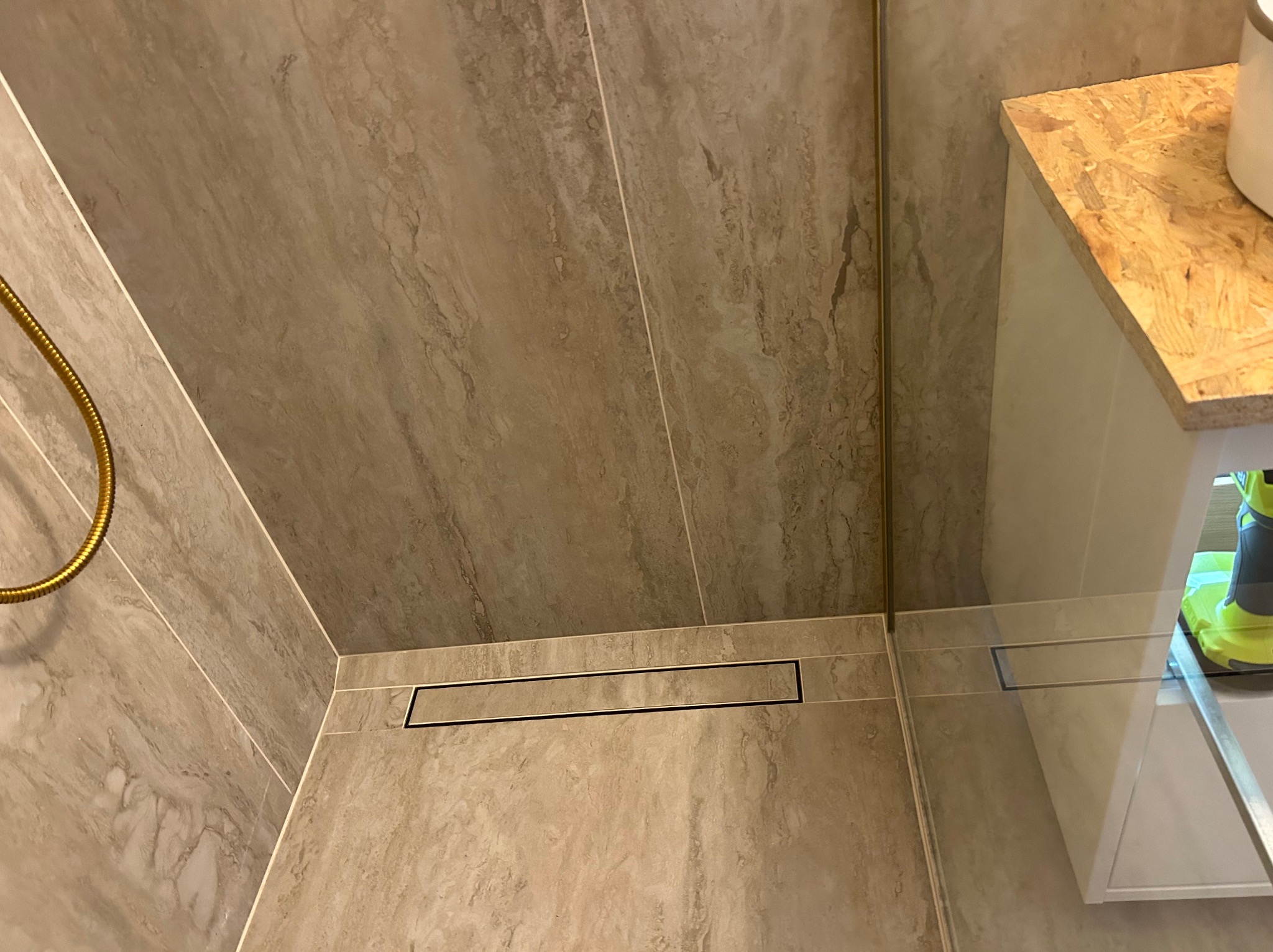Beżowe płytki imitujące marmur w kabinie prysznicowej z odpływem liniowym. Widoczny fragment słuchawki prysznicowej i szafki z blatem z płyty OSB.