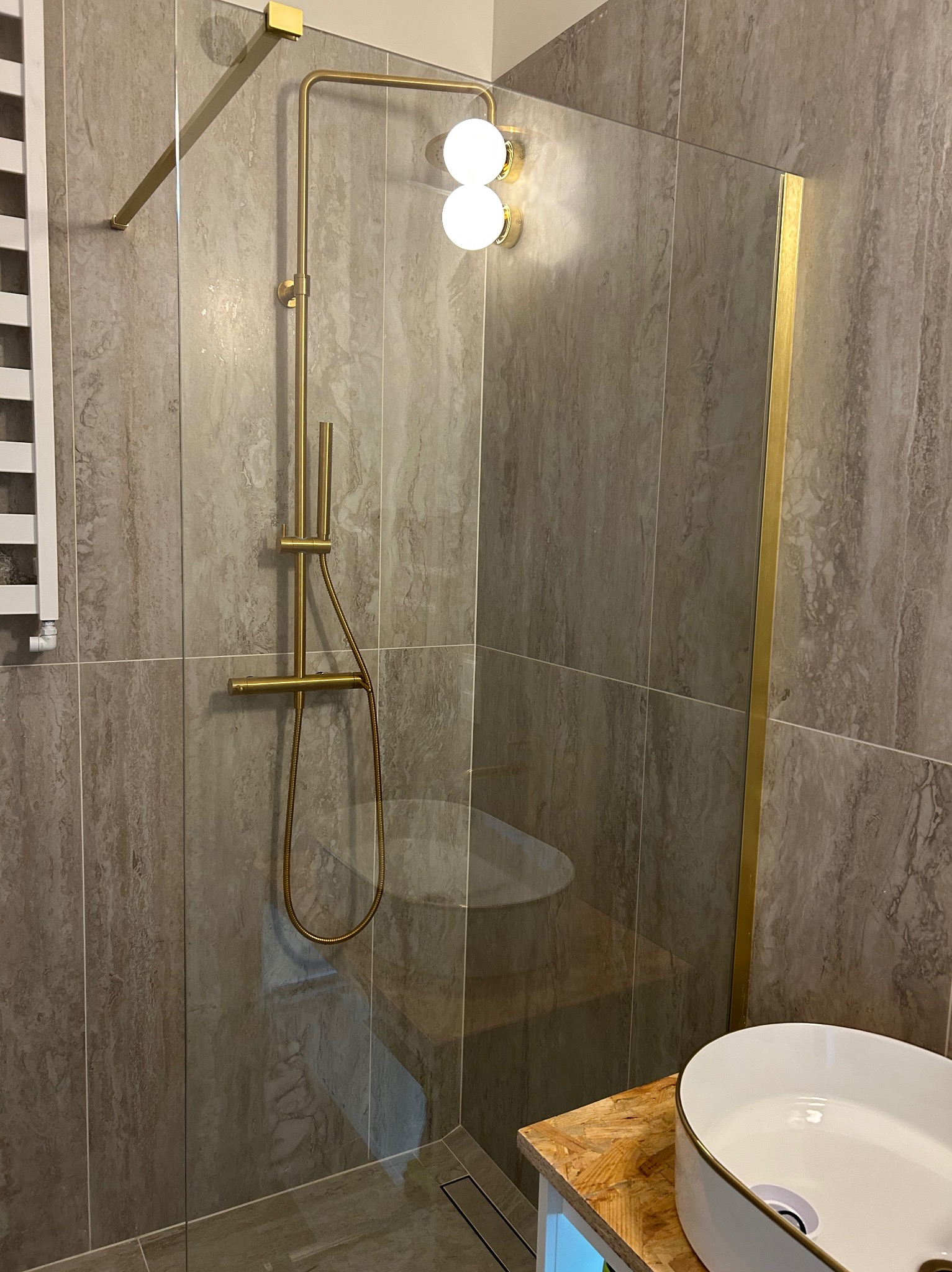 Wykończona łazienka z kabiną prysznicową typu walk-in, złotą armaturą i umywalką nablatową. Płytki imitujące kamień na ścianach i podłodze. Minimalistyczny design.
