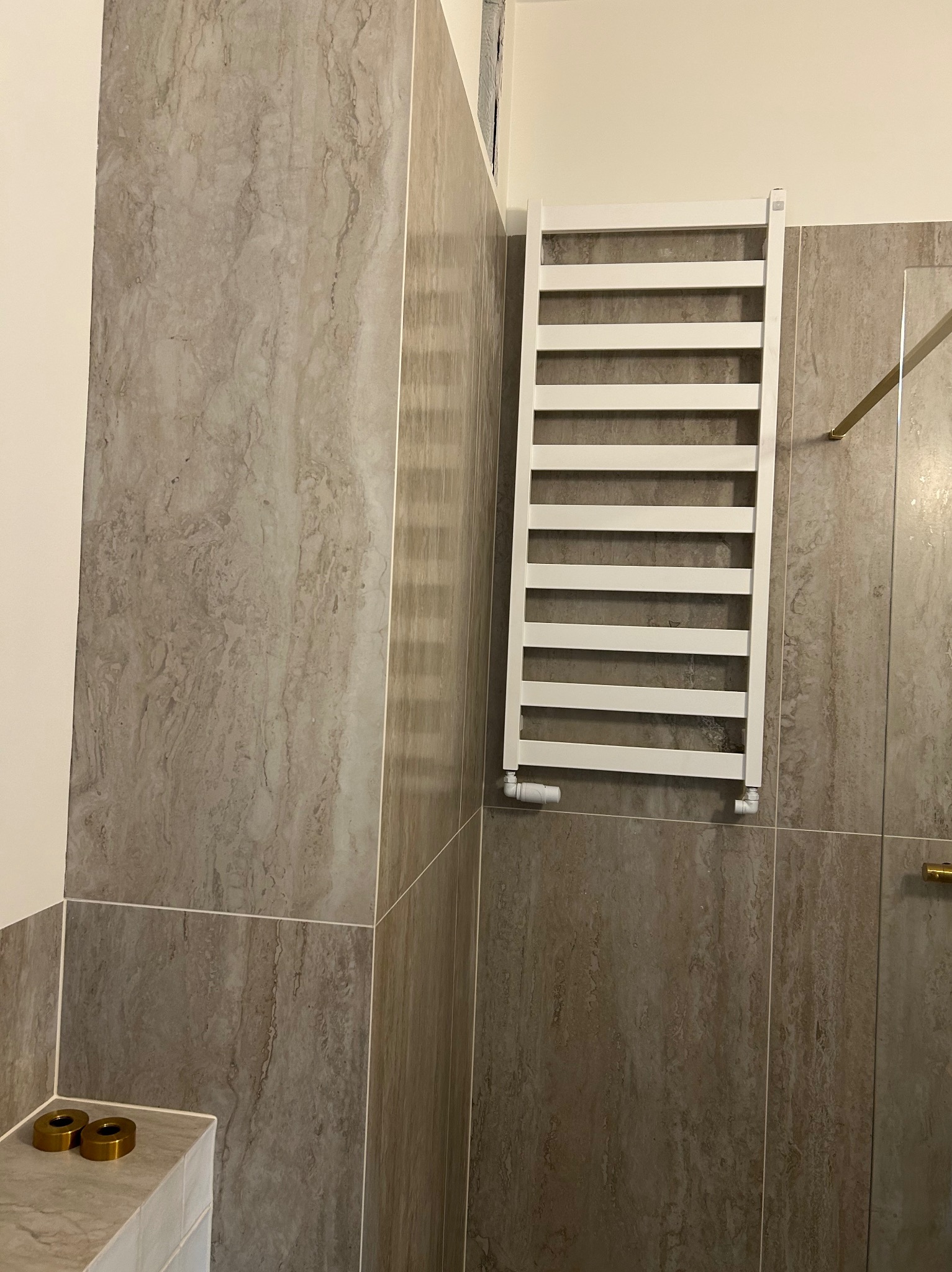 Wykończona łazienka z grzejnikiem drabinkowym na ścianie z płytek imitujących marmur. Widoczne złote elementy armatury i fragment kabiny prysznicowej.