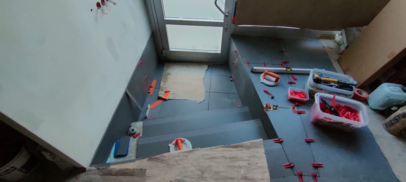 Układanie ciemnych płytek na schodach i podłodze, widoczne narzędzia glazurnicze i poziomica. Prace w toku, płytki zabezpieczone klipsami. Ujęcie z góry.