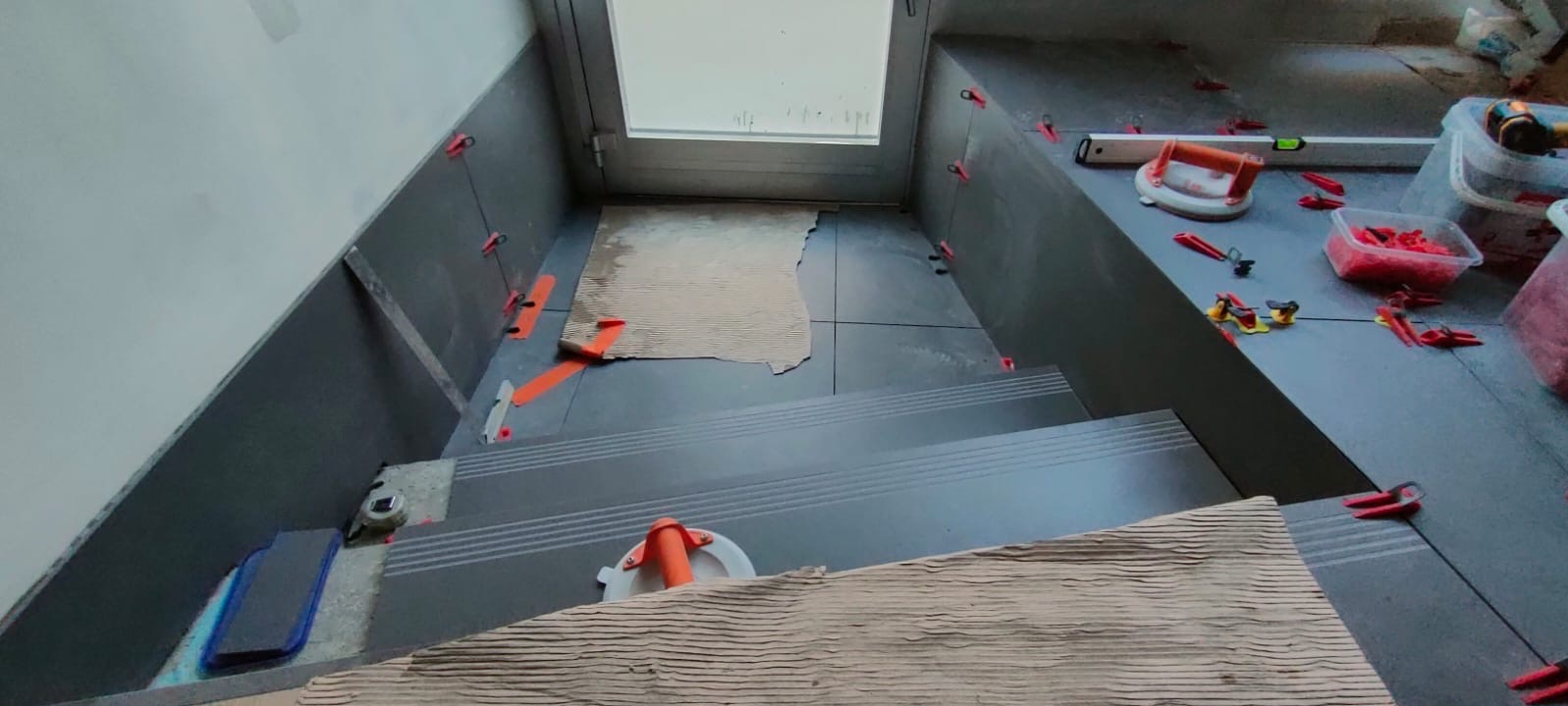 Układanie ciemnych płytek na schodach z użyciem systemu poziomowania. Widoczne narzędzia glazurnicze i fragmenty kartonu ochronnego. Praca w toku.