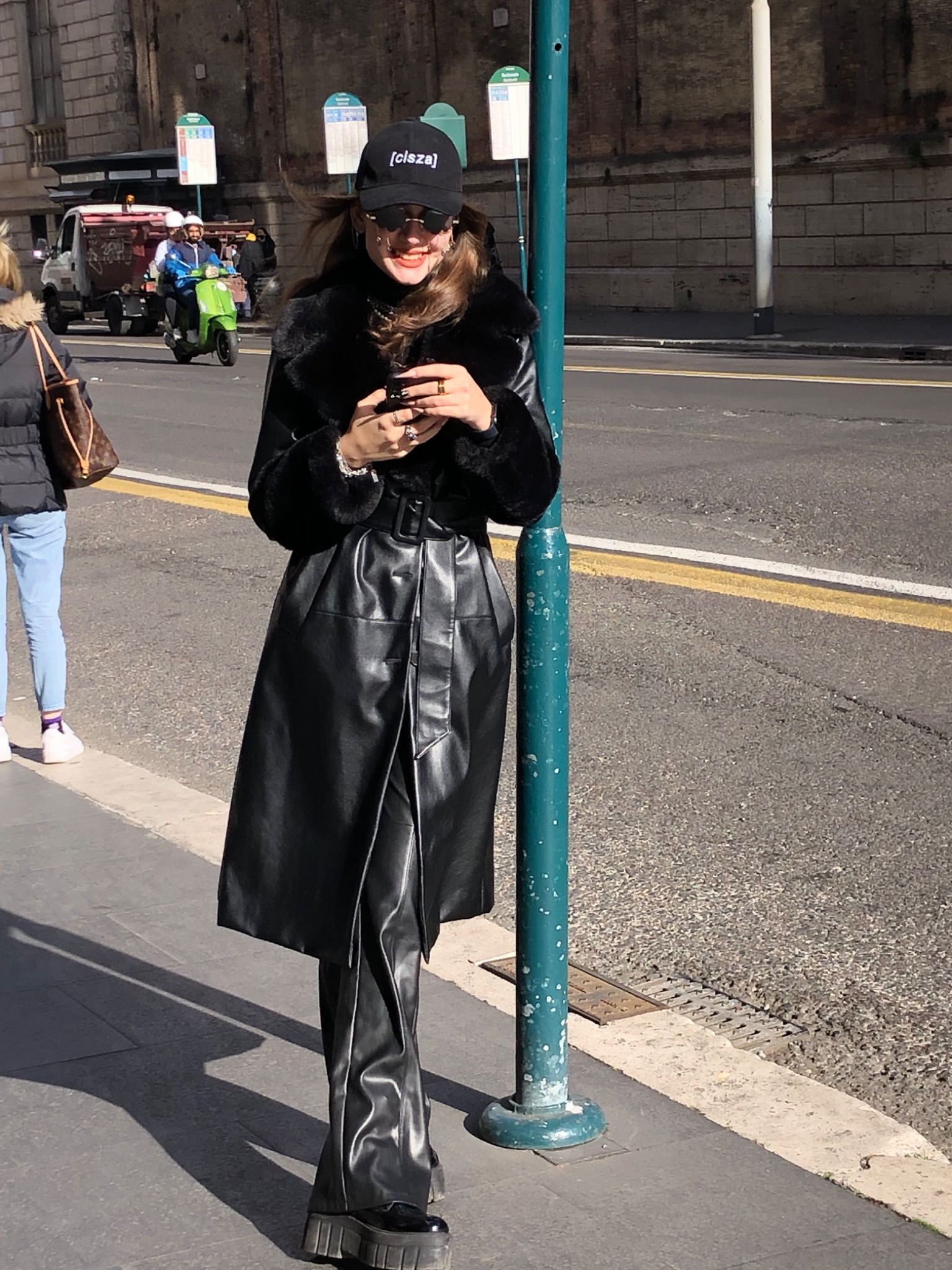 Uśmiechnięta kobieta w czarnym płaszczu ze sztucznej skóry i czapce z daszkiem, stojąca na chodniku obok zielonego słupka, trzyma telefon w dłoniach.