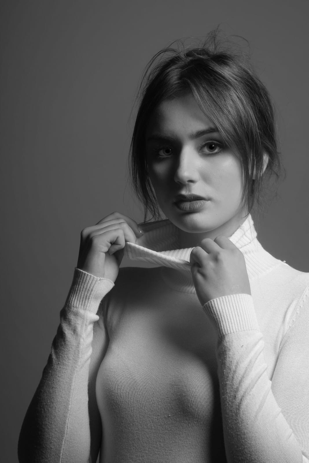 Portret młodej kobiety w białym golfie, czarno-białe zdjęcie z delikatnym oświetleniem, modelka trzyma kołnierz golfu uniesiony do góry.