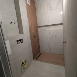 Wnętrze łazienki w trakcie remontu, widoczne płytki imitujące marmur na ścianach, drewnopodobne płytki w strefie prysznica, niezabudowany stelaż podtynkowy WC oraz rury kanalizacyjne wystające ze...