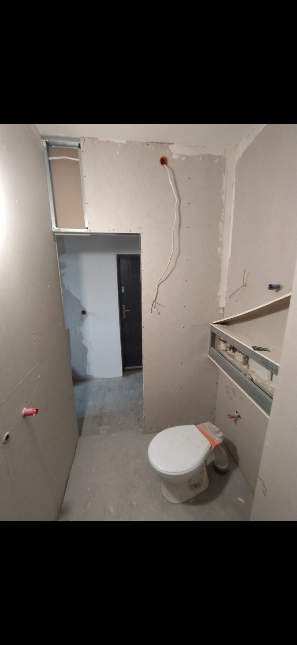 Wnętrze łazienki w trakcie remontu: ściany wyłożone płytami kartonowo-gipsowymi, widoczne profile metalowe, instalacja elektryczna i hydrauliczna w ścianach, zamontowana toaleta z zabezpieczeniem...