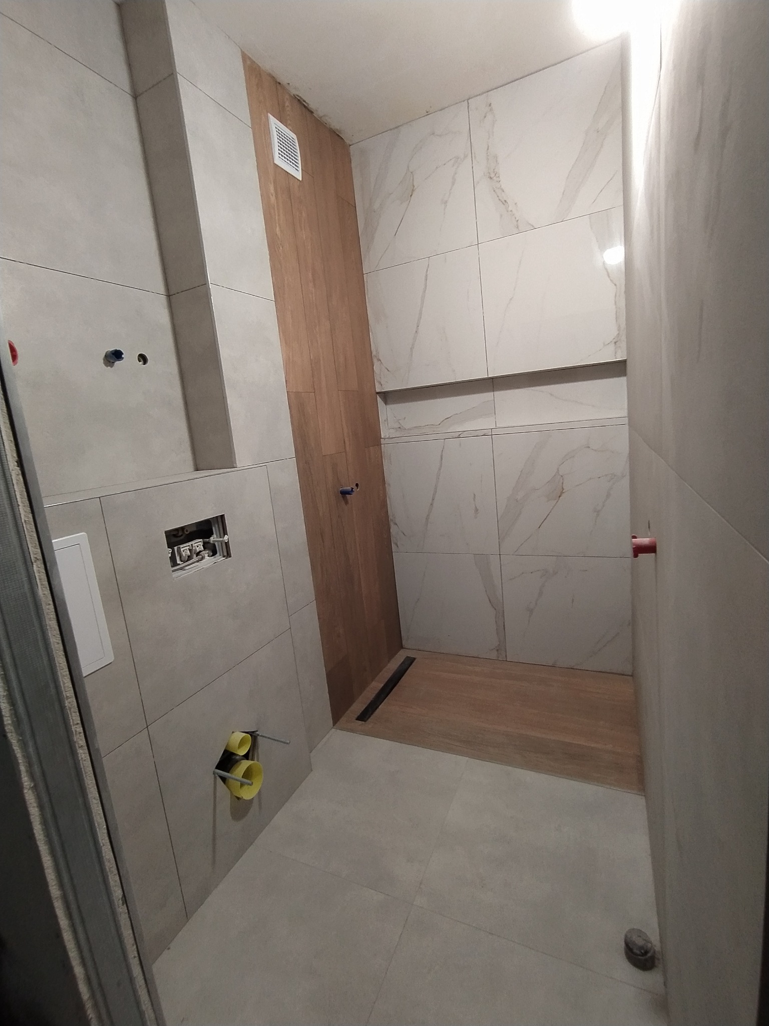 Wnętrze łazienki w trakcie remontu, widoczne płytki imitujące marmur na ścianach, drewnopodobne płytki w strefie prysznica, niezabudowany stelaż podtynkowy WC oraz rury kanalizacyjne wystające ze...