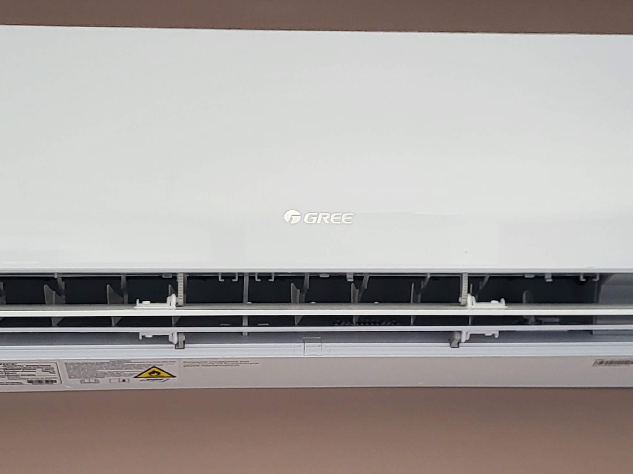 Biała jednostka wewnętrzna klimatyzatora marki Gree zamontowana na ścianie, widoczne żaluzje i naklejka ostrzegawcza.