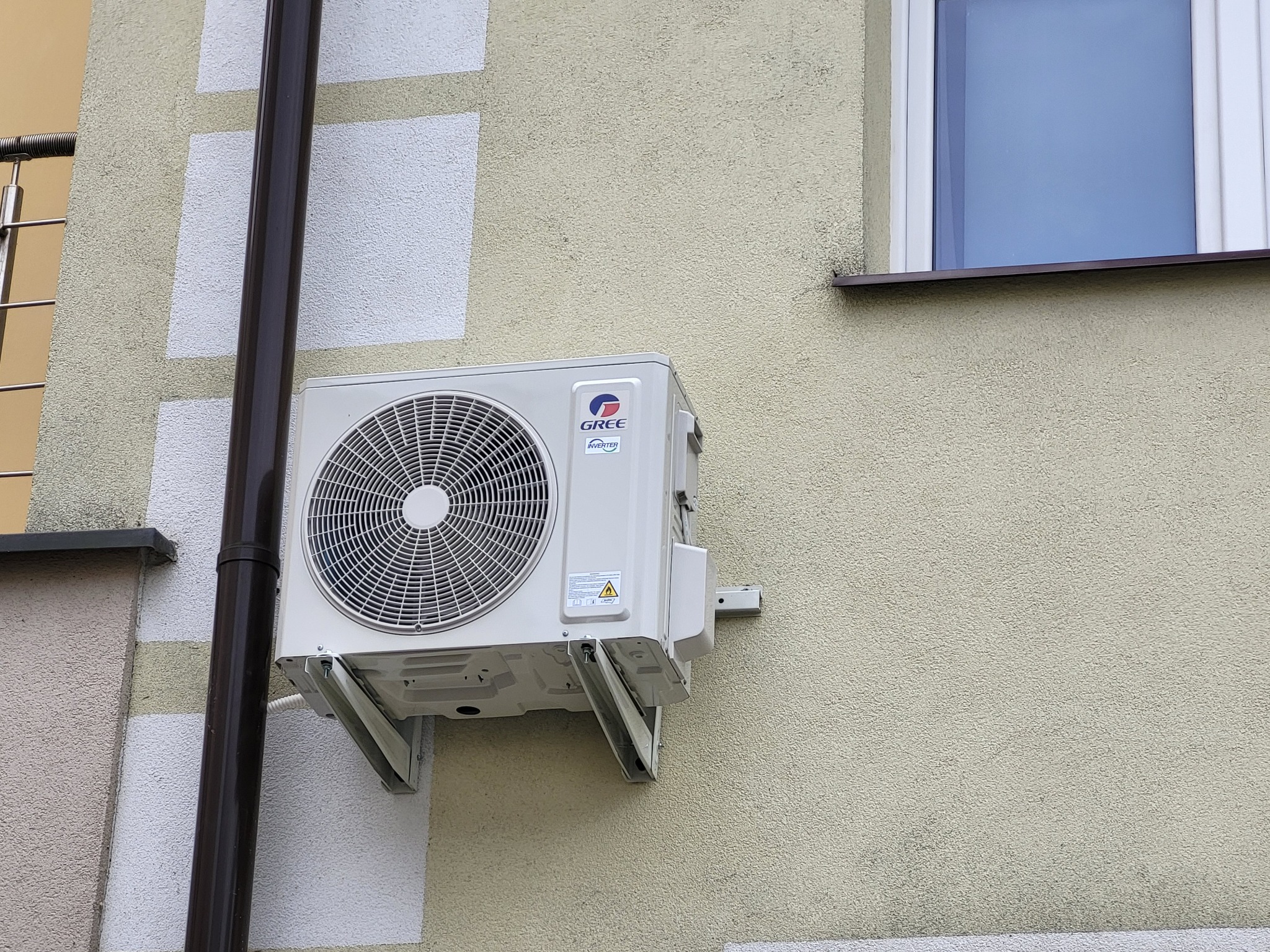 Zewnętrzna jednostka klimatyzacji marki Gree zamontowana na elewacji budynku z oknem, widoczna rura spustowa.