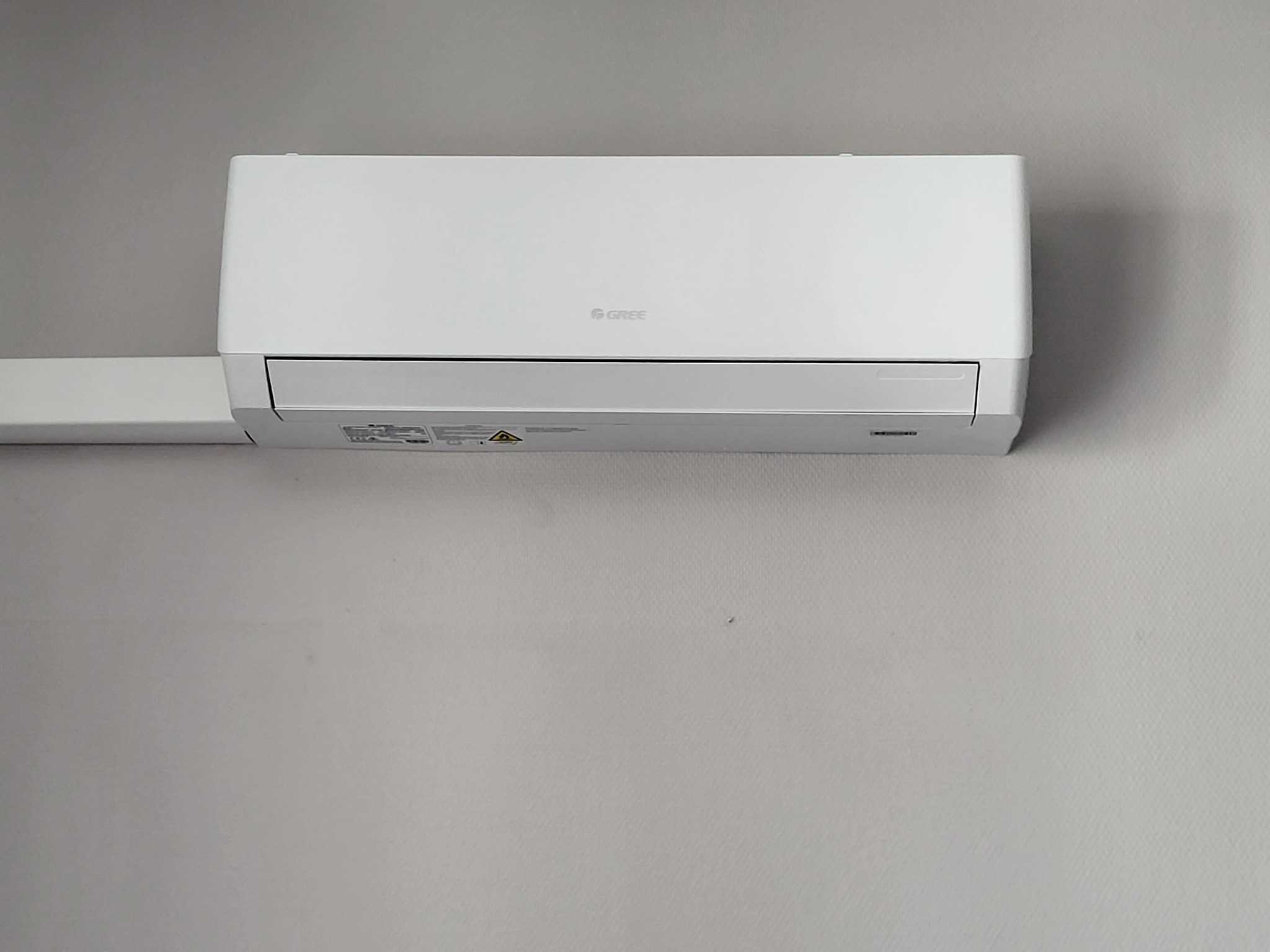 Biały klimatyzator marki Gree zamontowany na szarej ścianie, widoczny fragment instalacji elektrycznej.