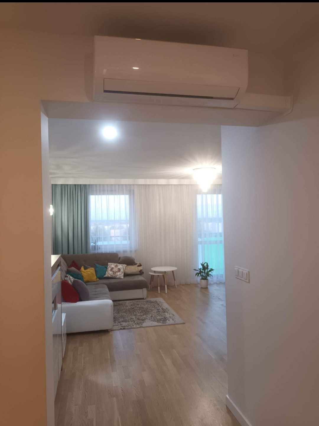 Nowoczesny salon z zamontowanym białym klimatyzatorem ściennym, widok z korytarza, jasne wnętrze z drewnianą podłogą i szarym dywanem.