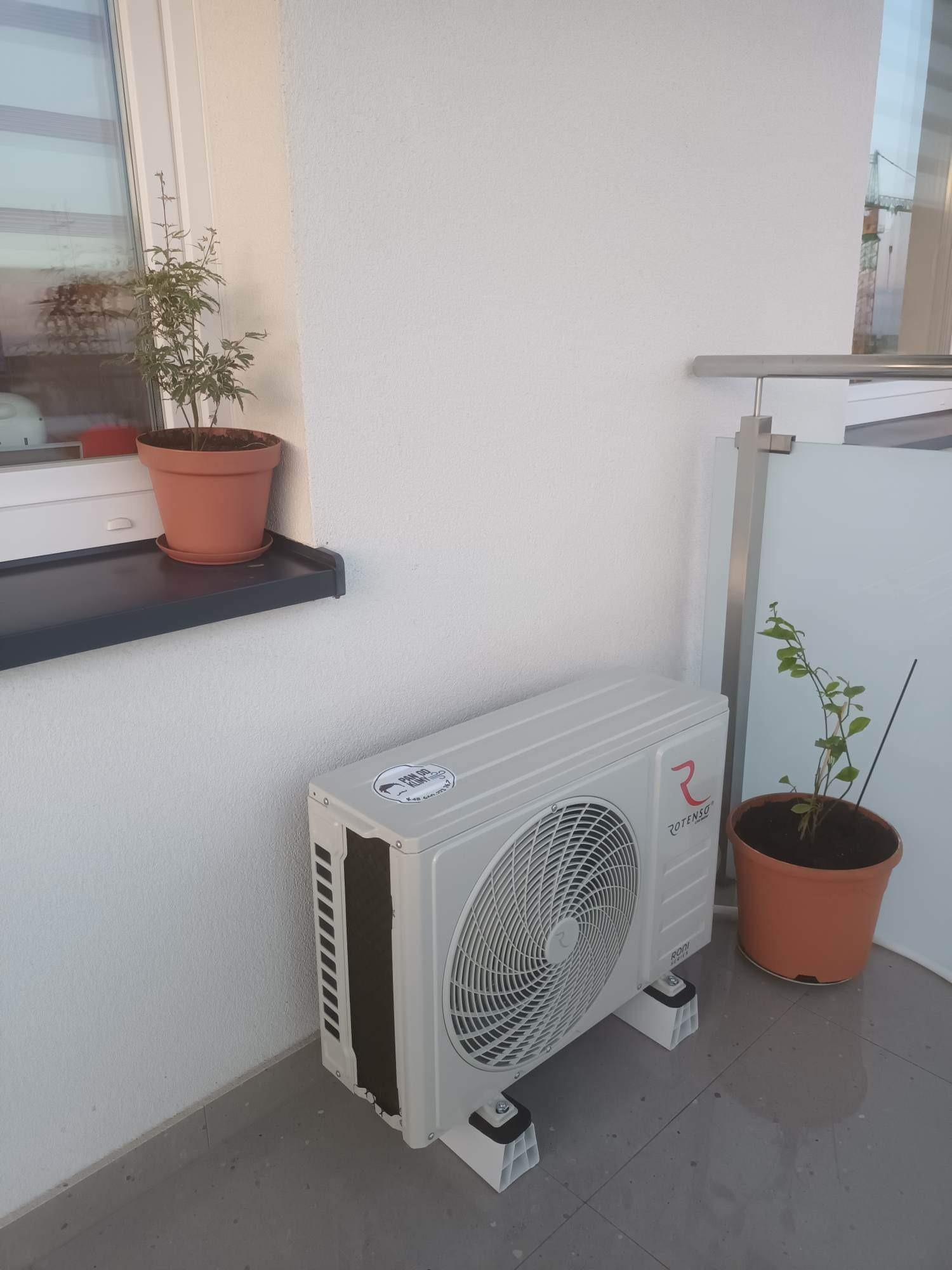 Zewnętrzna jednostka klimatyzacji Rotenso na balkonie, na nóżkach, obok doniczka z rośliną, w tle widoczny żuraw budowlany.