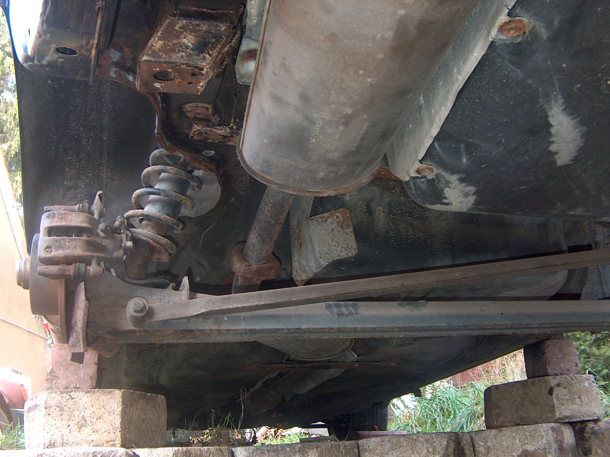 Podwozie samochodu z widocznymi elementami zawieszenia, układu wydechowego i korozją na metalowych częściach, podparte na betonowych blokach.