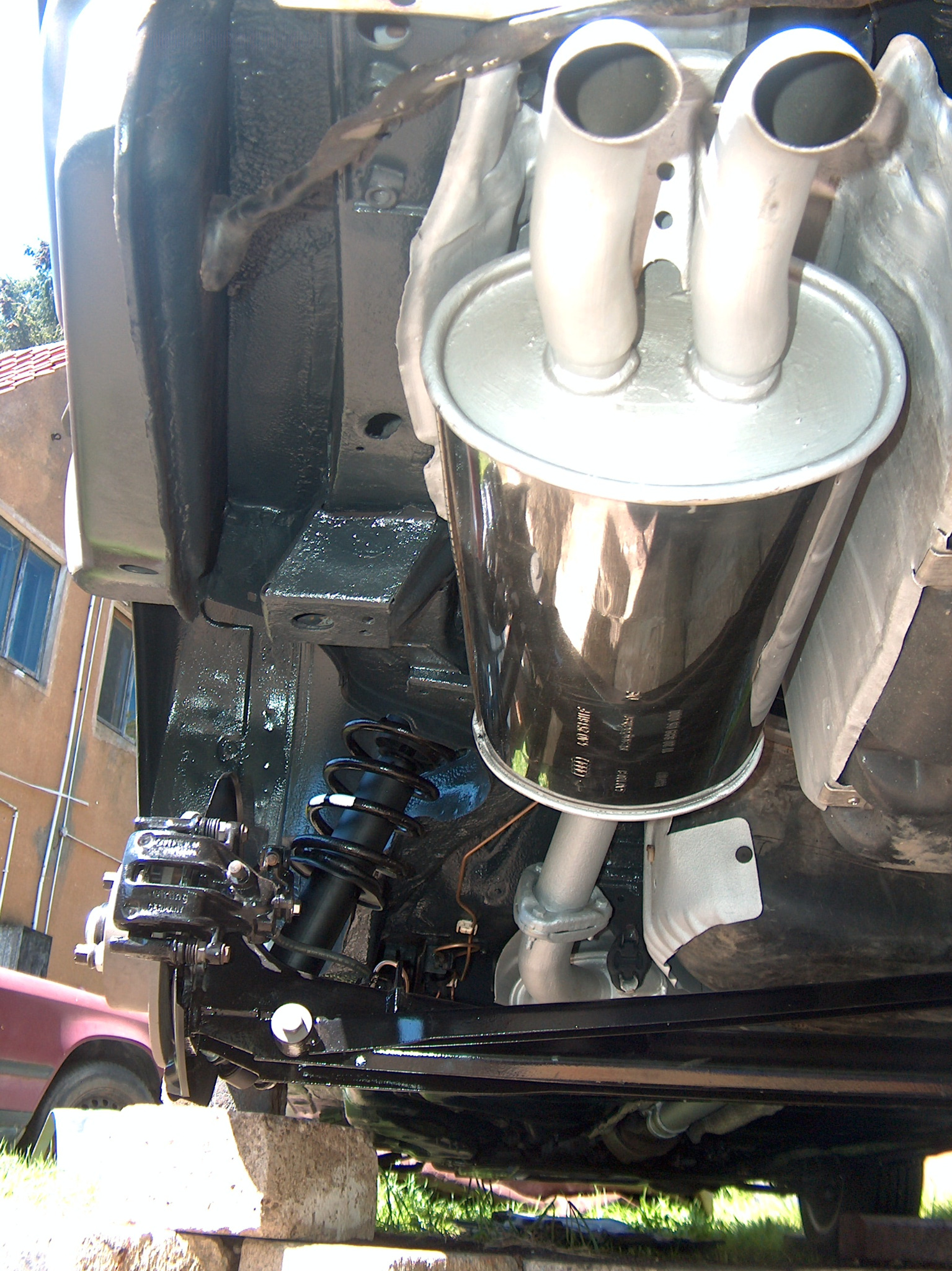 Podwozie auta po konserwacji: czarne elementy zawieszenia, srebrny tłumik z dwoma rurami. Widoczne sprężyny i fragment nadwozia, całość na betonowych blokach.