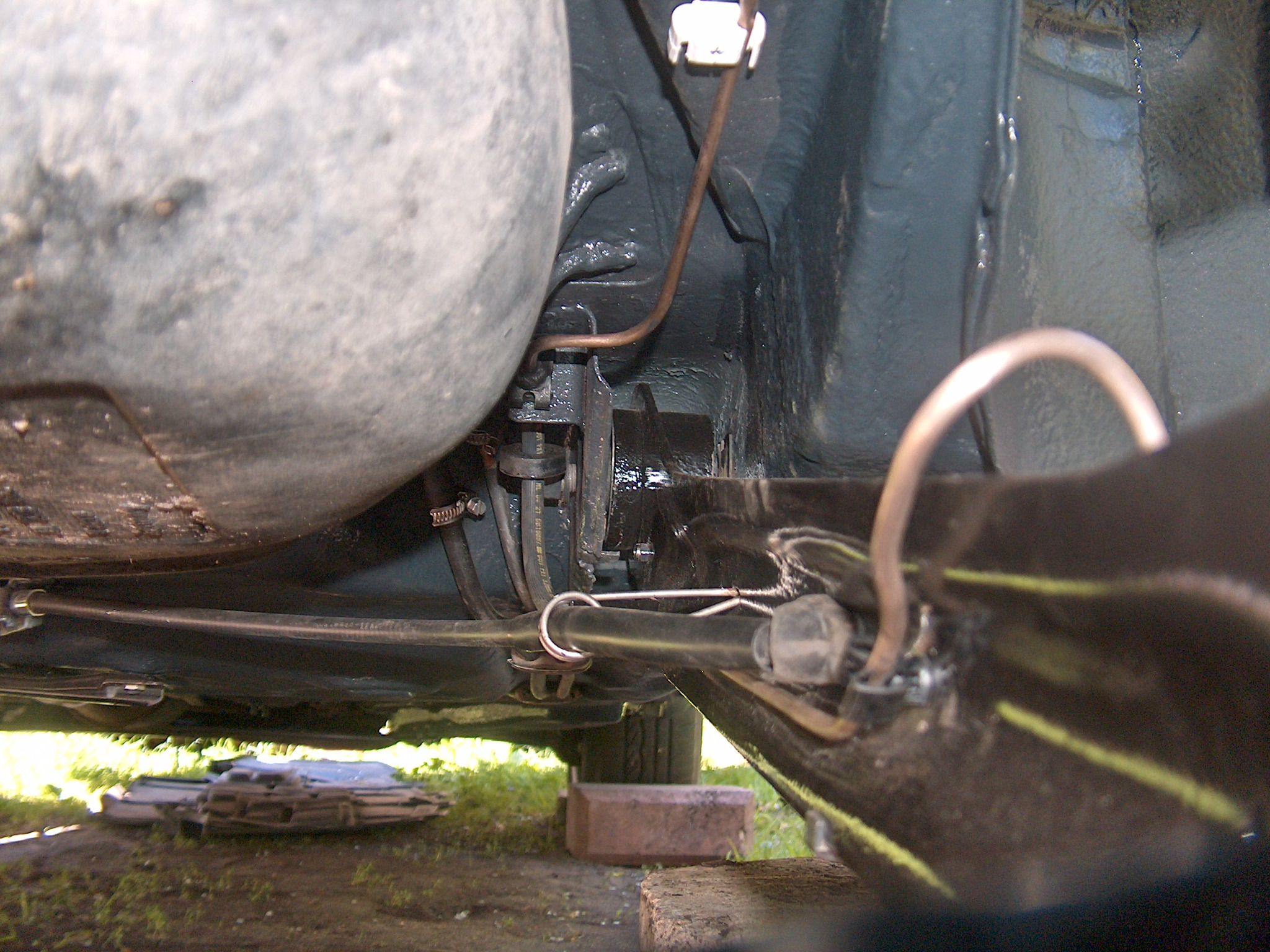 Spód pojazdu w trakcie konserwacji: widoczne elementy zawieszenia, przewody i podwozie zabezpieczone farbą antykorozyjną. Blok oporowy pod kołem, trawa w tle.