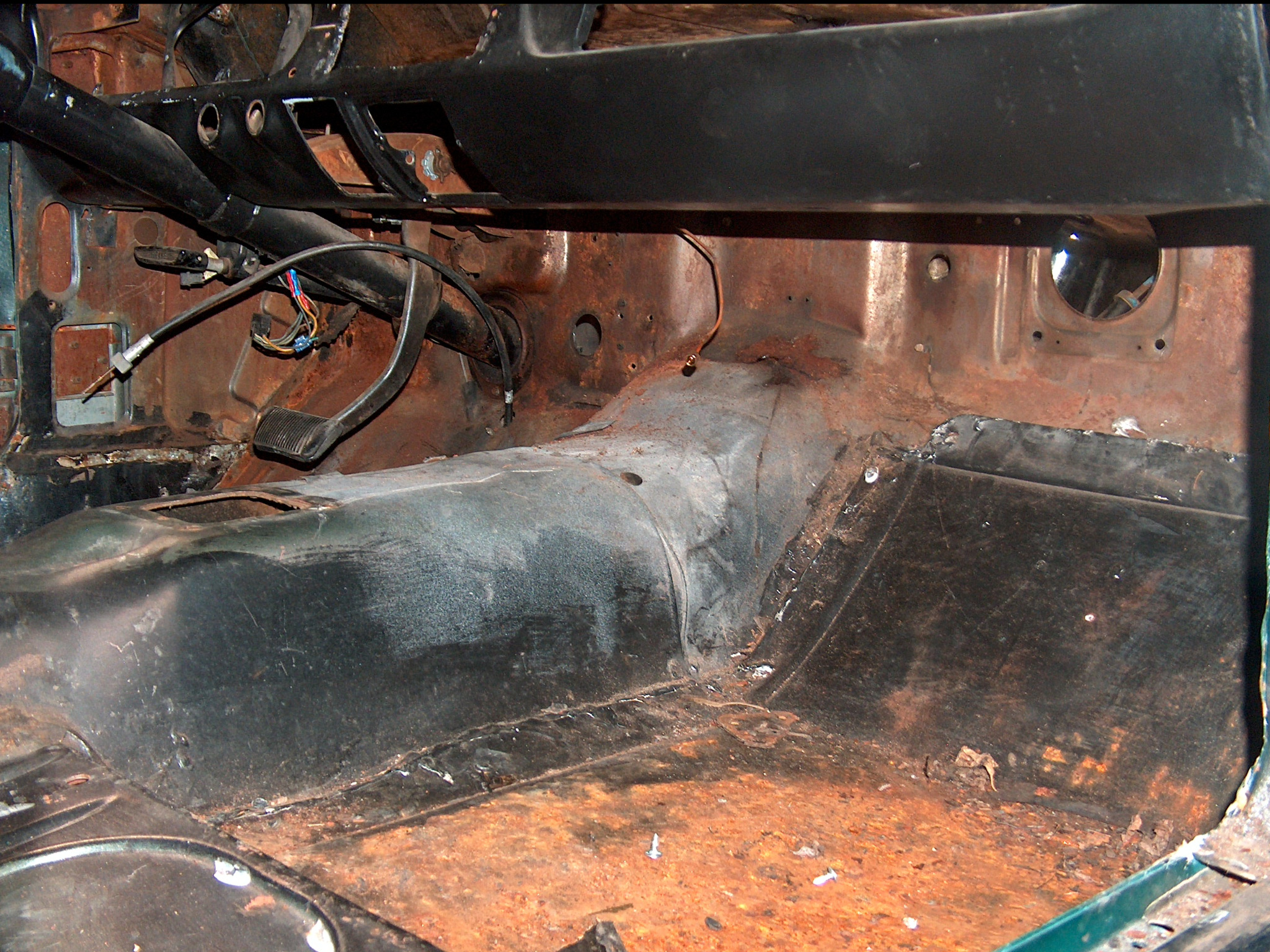 Wnętrze skorodowanego pojazdu przed renowacją. Widoczne elementy metalowe z rdzą, pedał hamulca, przewody. Perspektywa z wnętrza kabiny na podłogę i deskę rozdzielczą.