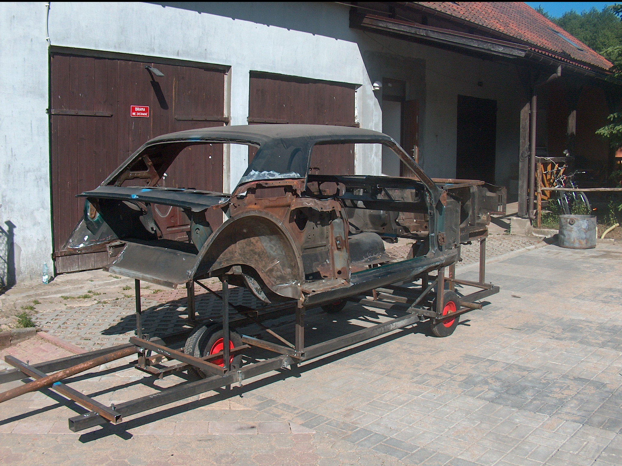 Karoseria klasycznego auta na metalowym wózku, widoczna rdza, przygotowana do renowacji na tle garażu z drewnianymi bramami i brukowanej nawierzchni.