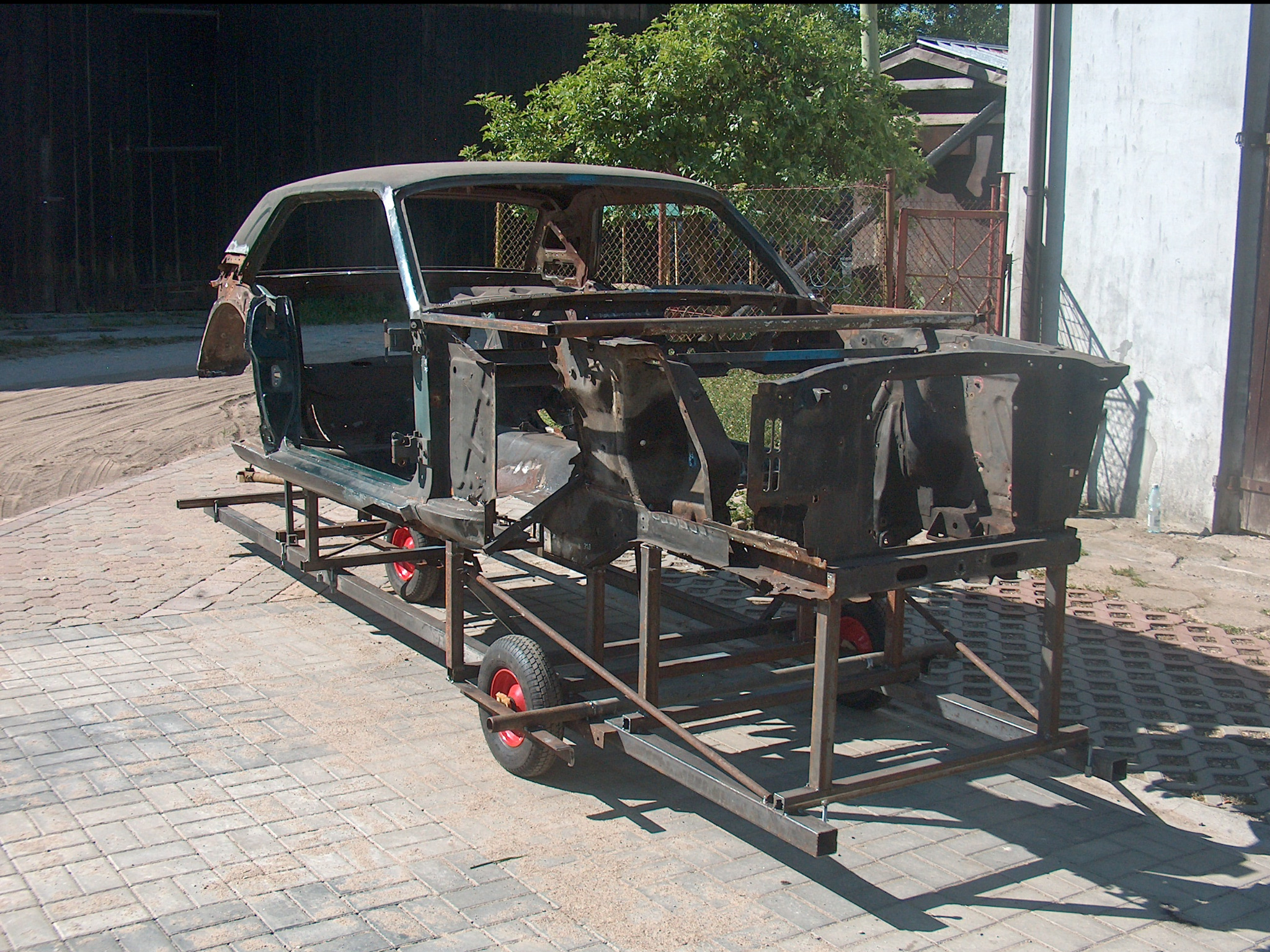 Karoseria klasycznego samochodu, częściowo zdemontowana, umieszczona na metalowej platformie z czerwonymi kołami. Prace przygotowawcze do renowacji w plenerze.